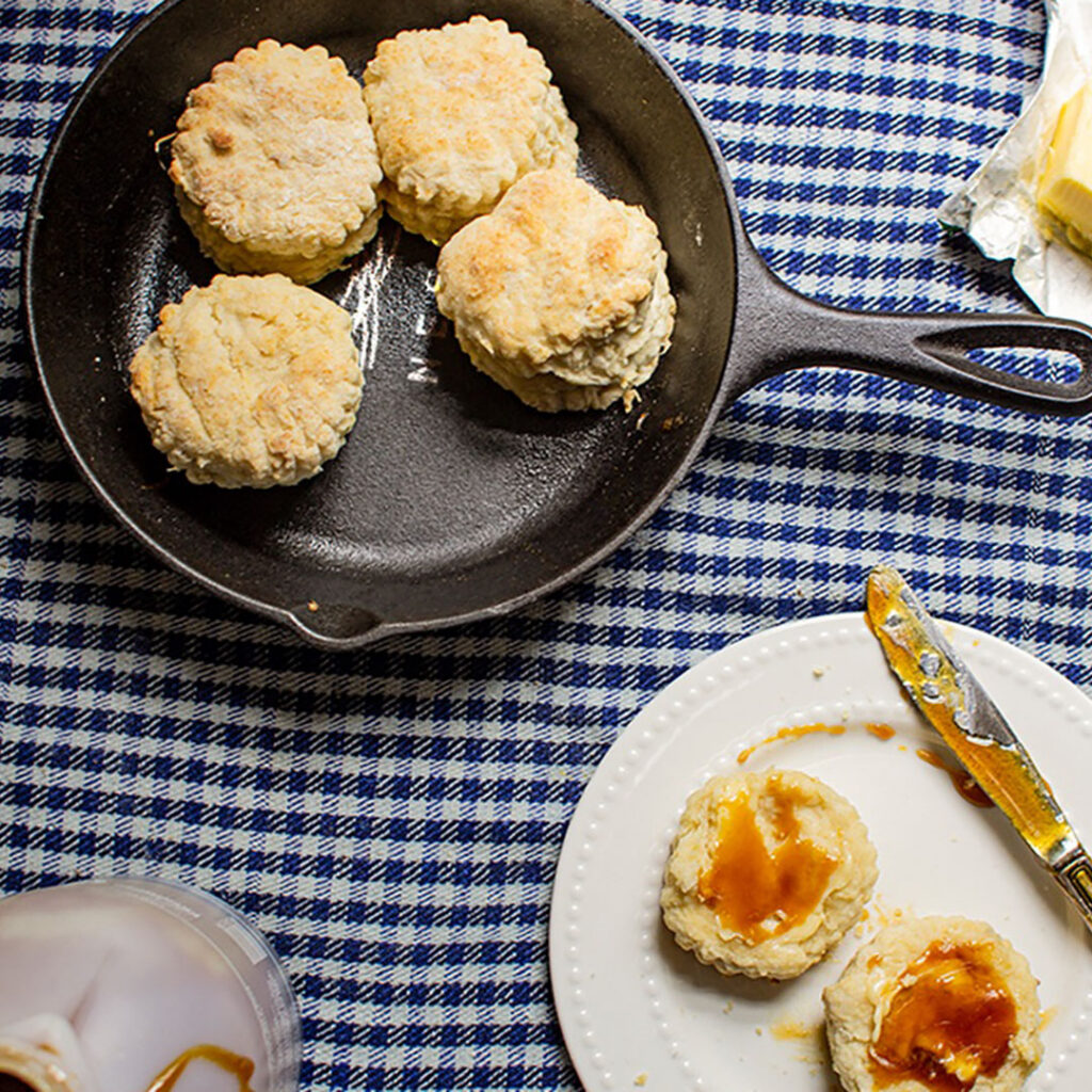 Four golden biscuits in a black cast iron skillet on a blue-and-white cloth, with butter and syrup served on a nearby plate.