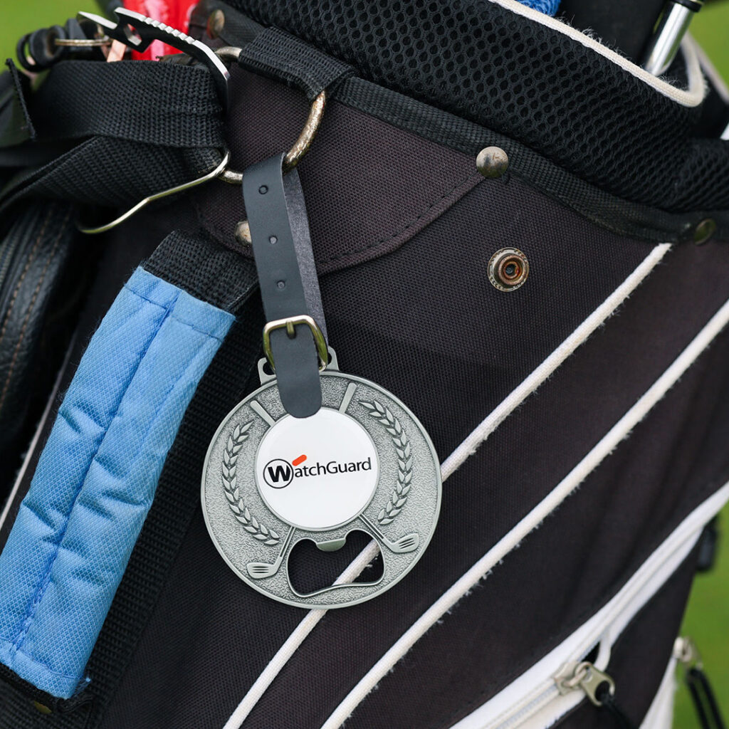 Custom WatchGuard golf bag tag attached to a black golf bag, featuring a round metal tag with a built‑in bottle opener.