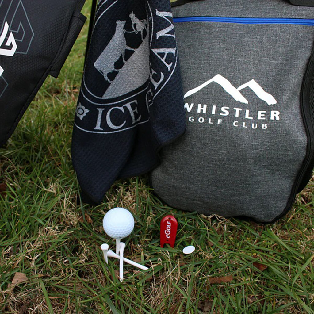 Branded golf kit on grass with a Whistler Golf Club drawstring bag, golf towel, ball, tees, and a red wooden tee.