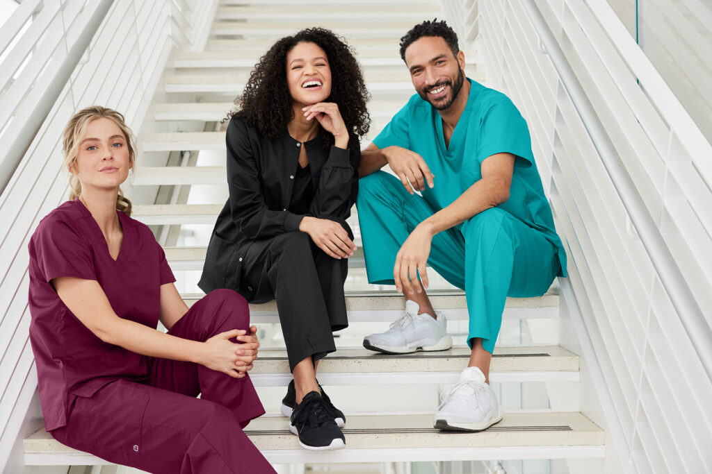 Three people wearing medical scrubs seated on a white staircase inside a bright building.