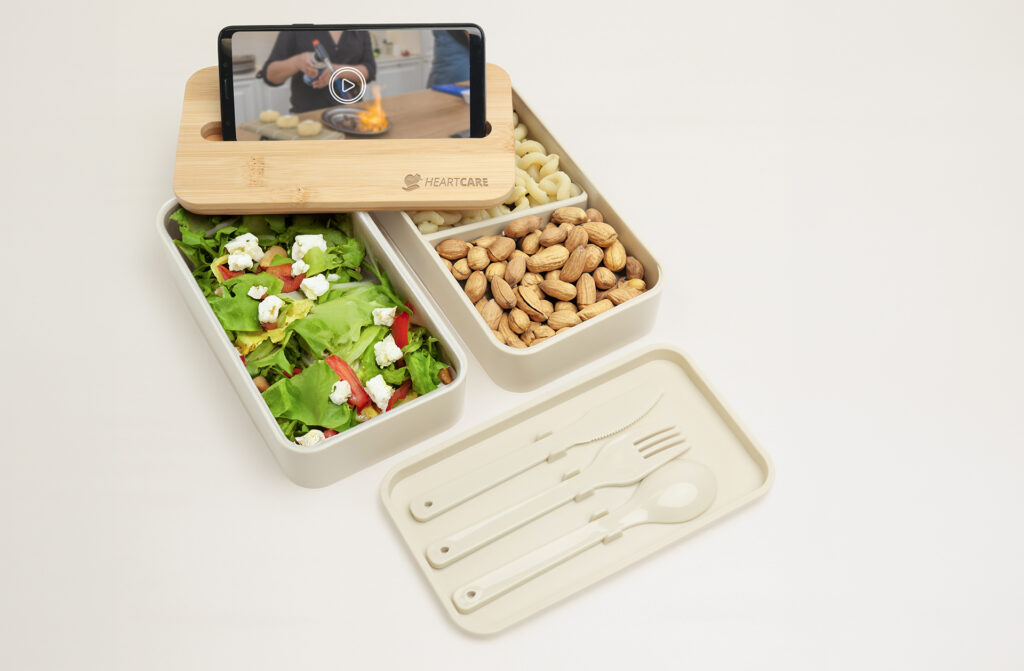 Bento-style lunch container with a salad, nuts, and a wooden phone stand holding a smartphone, with reusable cutlery beside it.