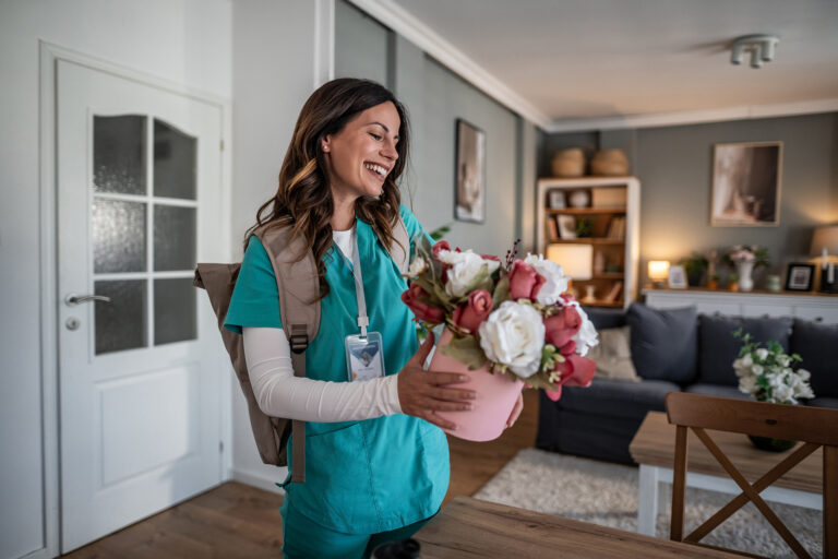Person in teal medical scrubs with an ID badge placing a pink flower arrangement on a table in a living room.