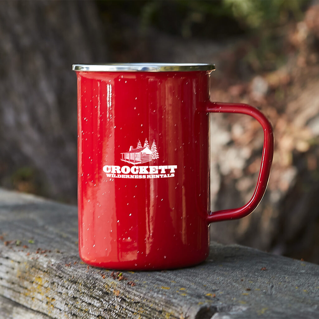 Red enamel camp mug with “Crockett Wilderness Rentals” logo resting on a wooden fence outdoors.
