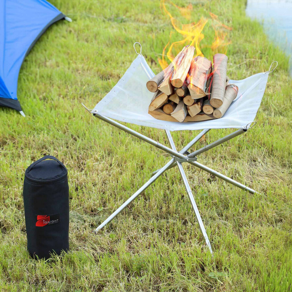 Portable camping fire pit holding stacked firewood with active flames on a grassy campsite near a blue tent.