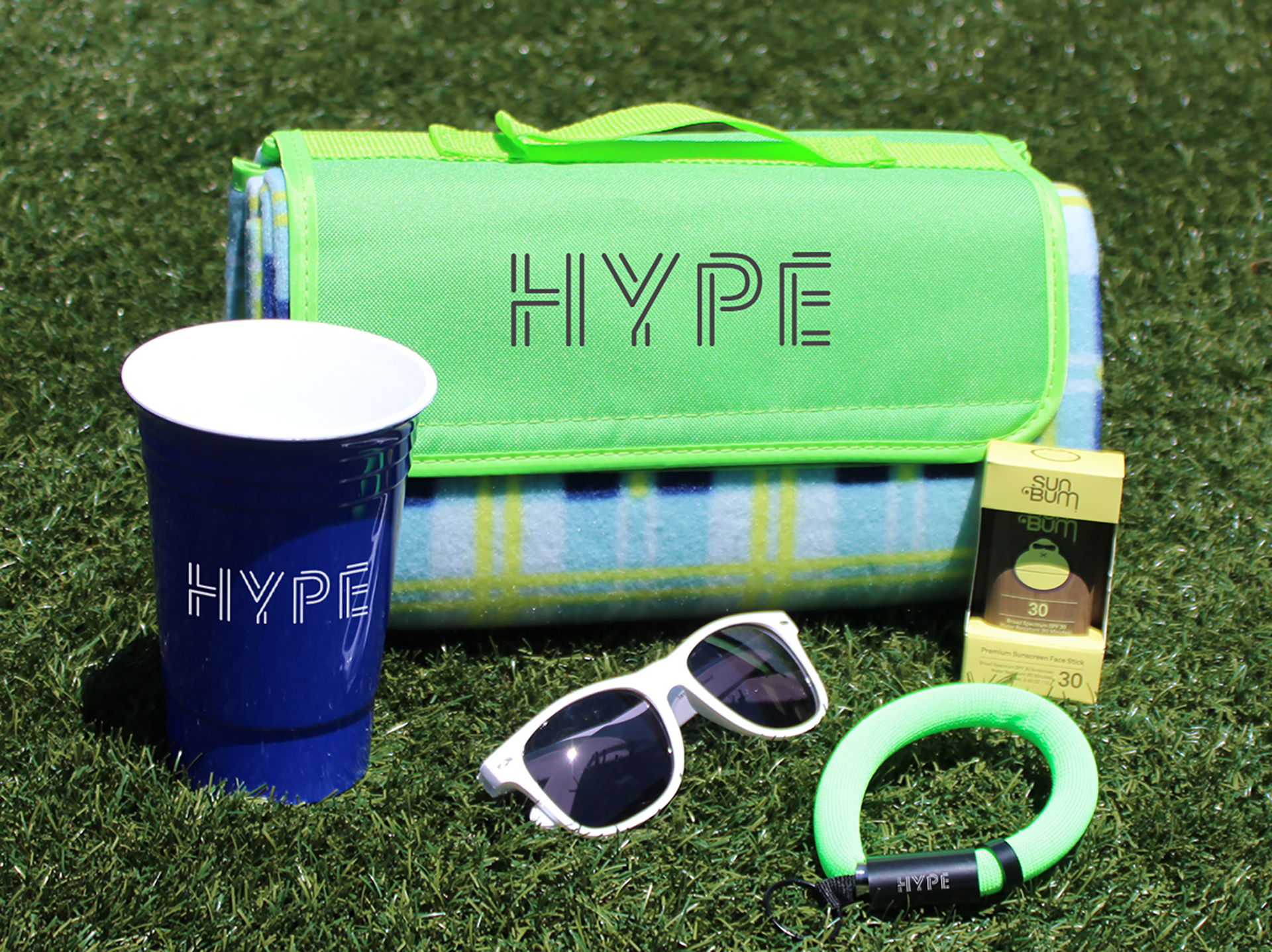 Branded outdoor merchandise including picnic blanket, reusable cup, sunglasses, sunscreen, and wristband arranged on grass.