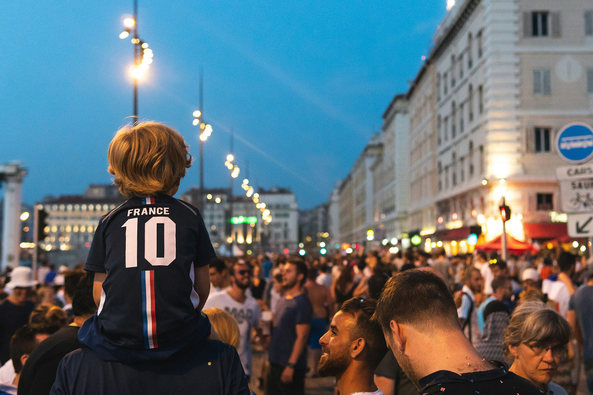 Soccer fans gathered on a city street at dusk, with a child on shoulders wearing a France number 10 jersey.