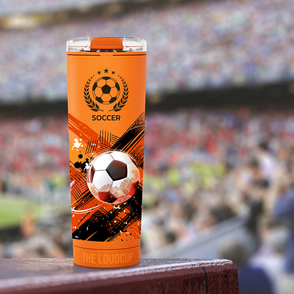 Soccer-themed orange drink cup with a ball design, photographed in a stadium setting with spectators in the background.