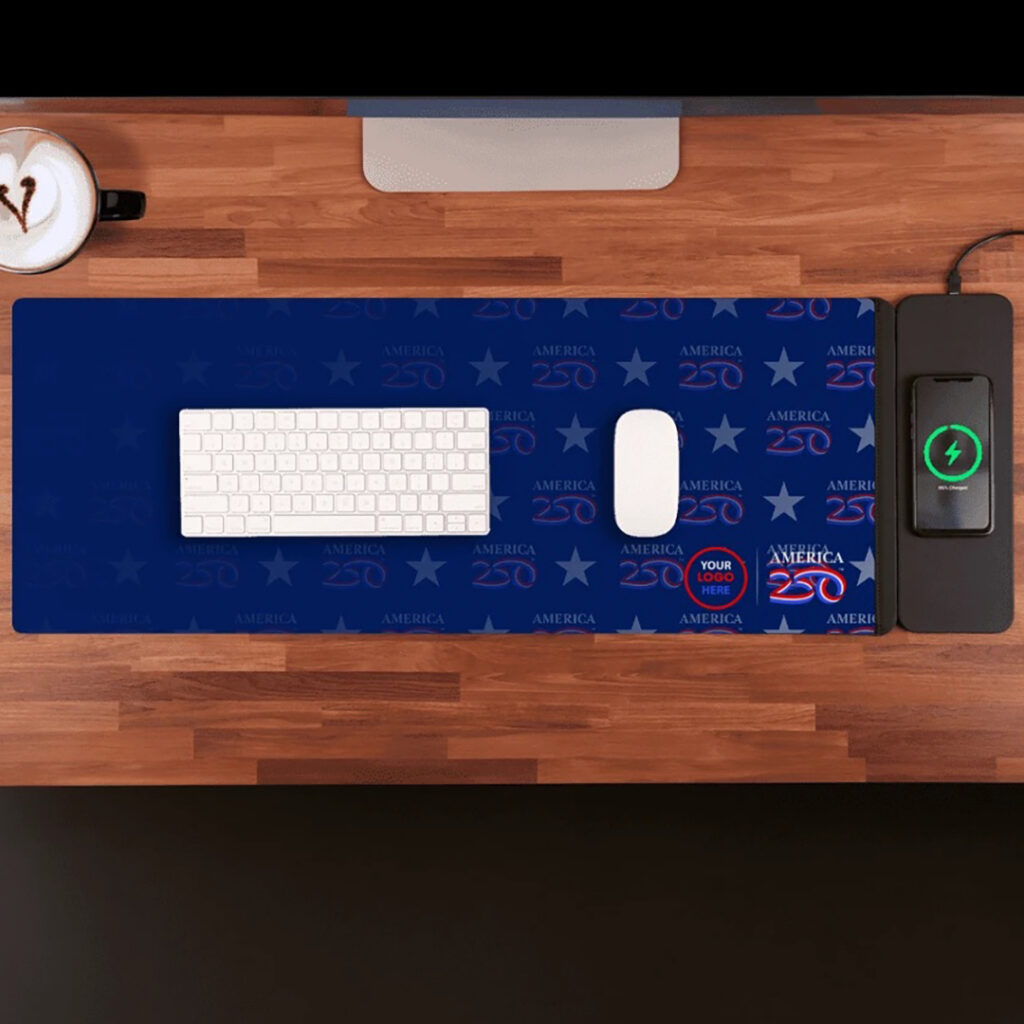 Top-down view of a wooden desk with a blue “America 250” branded desk mat, white keyboard and mouse, smartphone on a wireless charger, and a cup of coffee.