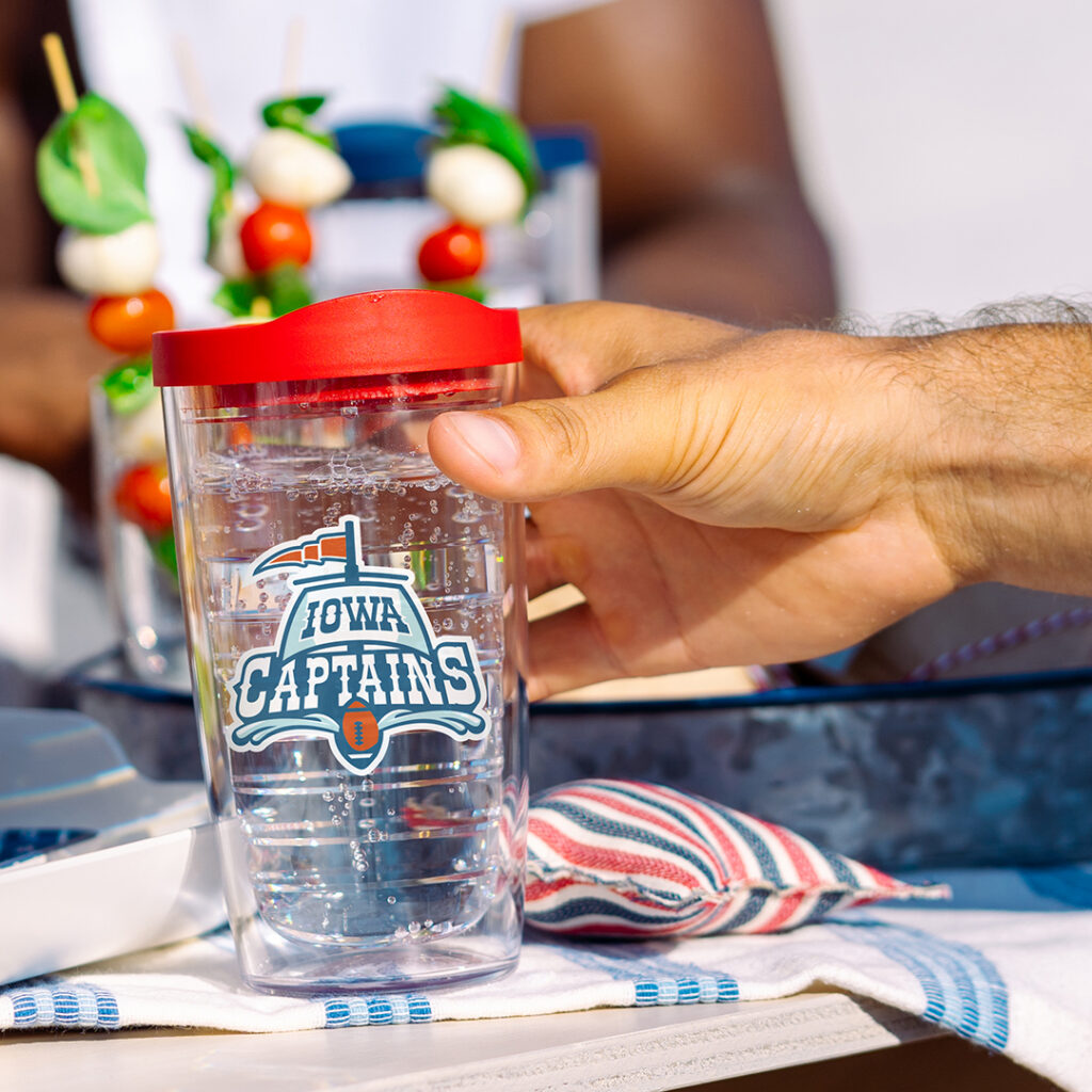 Hand holding a clear tumbler with a red lid and “Iowa Captains” logo, filled with sparkling water, with skewered snacks and a picnic-style setting in the background.