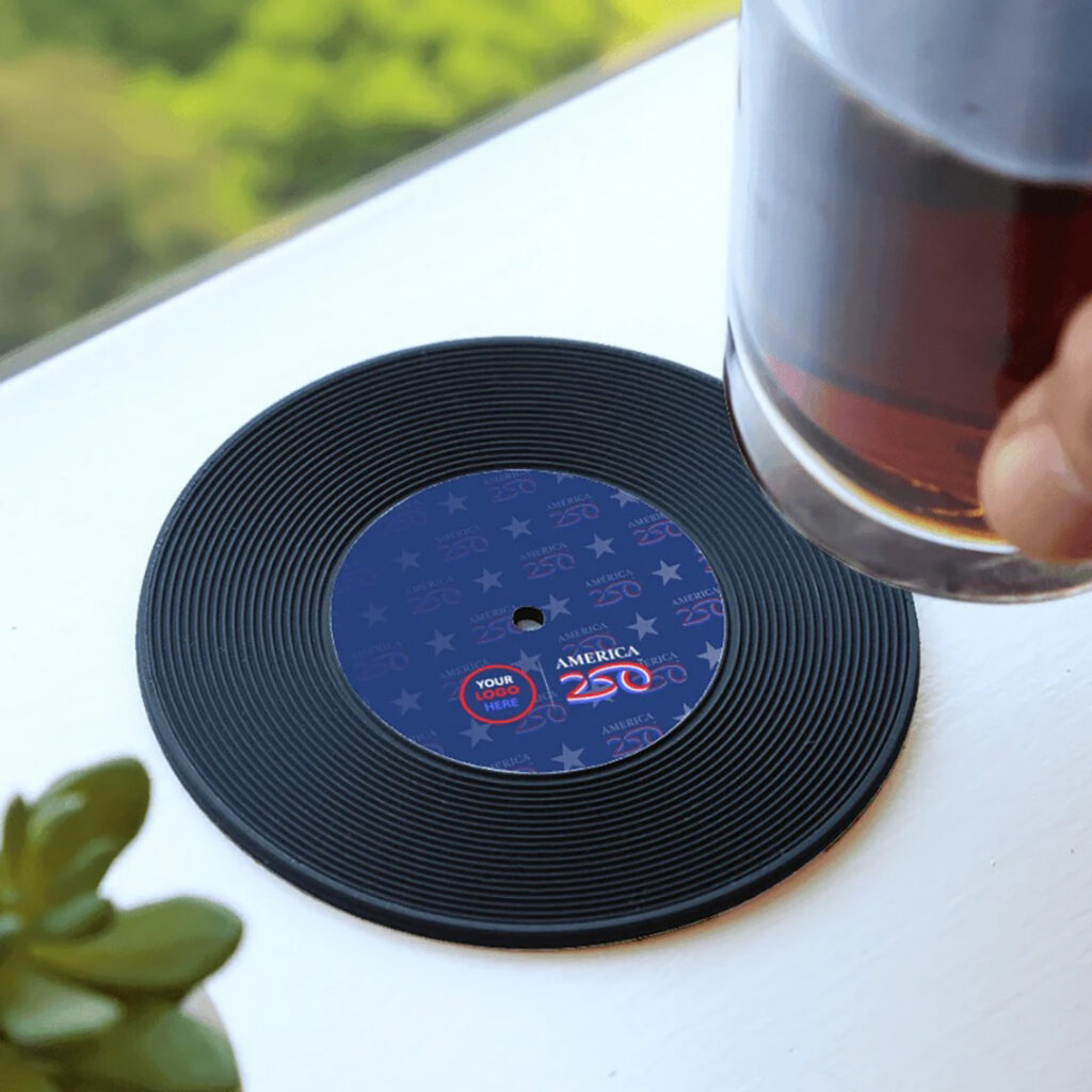 Glass of iced drink being placed on a black record-style coaster with a blue “America 250” design and customizable logo area on an outdoor table.
