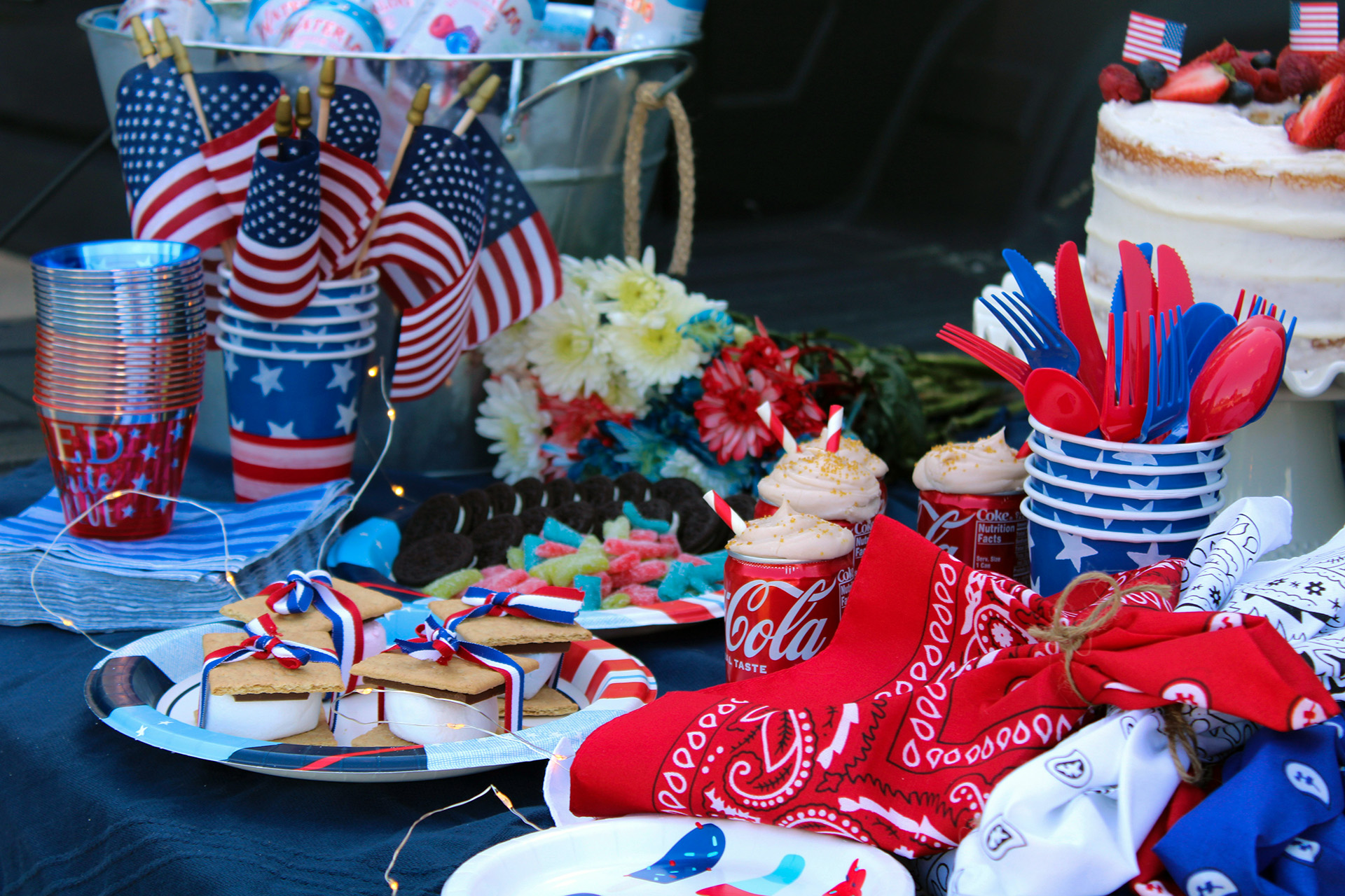 Festive red, white, and blue party table with American flags, patriotic tableware, cupcakes, cookies tied with ribbons, and a decorated cake, arranged for a Fourth of July celebration outdoors.