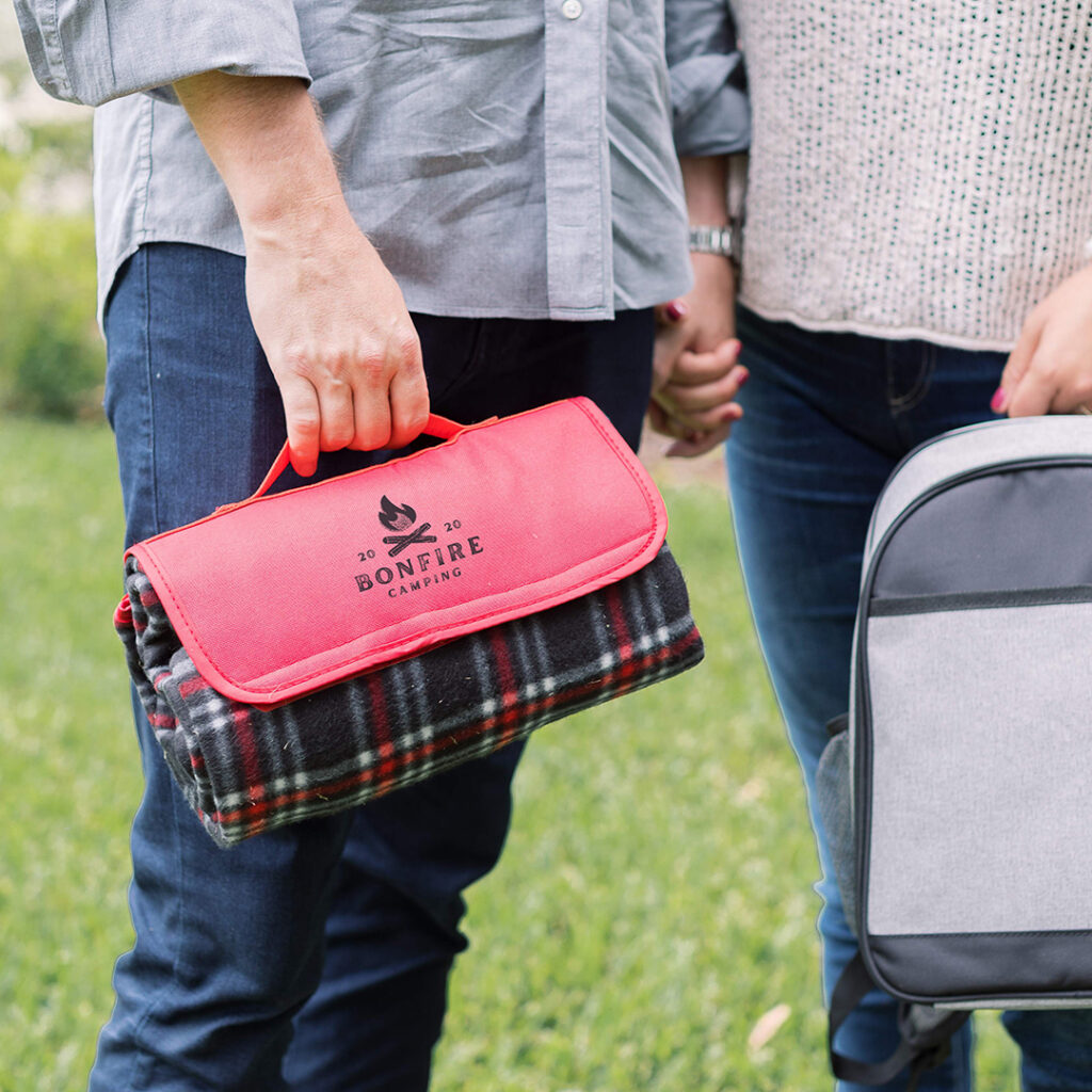Close-up of two people walking outdoors holding hands, one carrying a rolled plaid picnic blanket in a red “Bonfire Camping” carrier and the other holding a gray cooler bag.