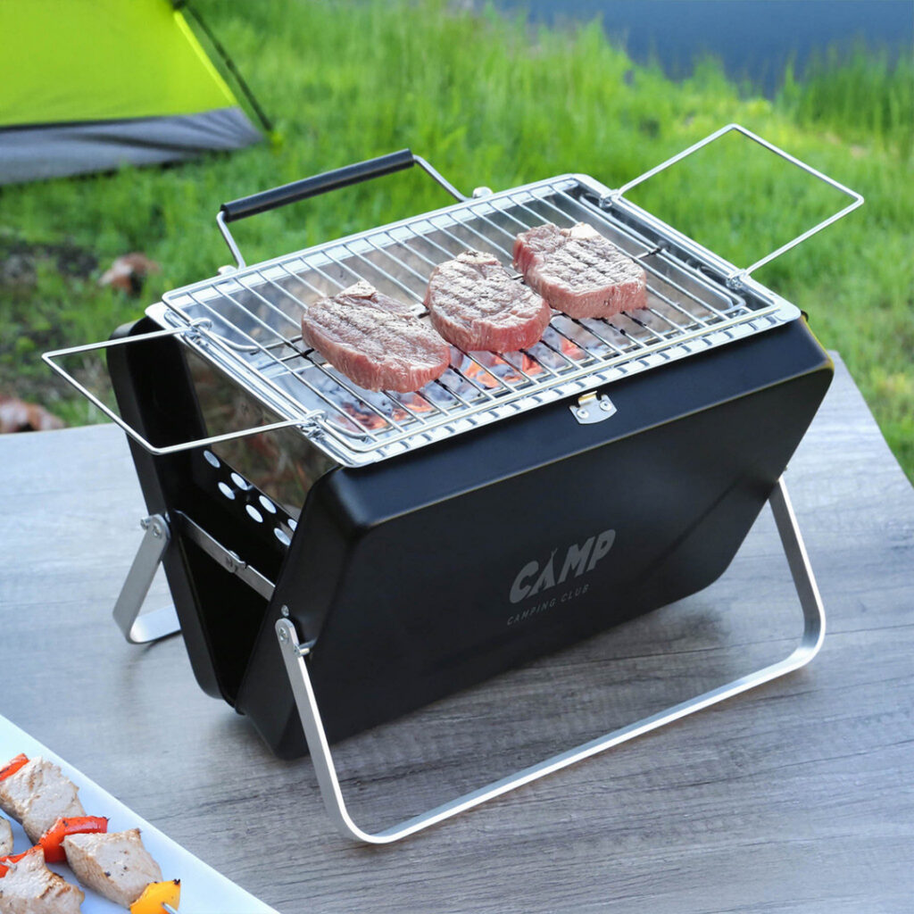 Portable tabletop charcoal grill with steaks cooking over hot coals, set outdoors on a wooden surface near a camping area with a tent in the background.