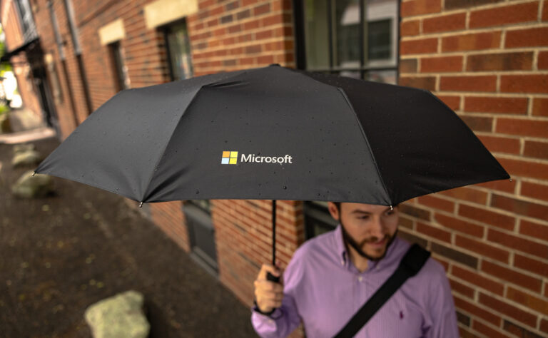 Person holding a black logo-printed umbrella with Microsoft branding on a rainy urban sidewalk