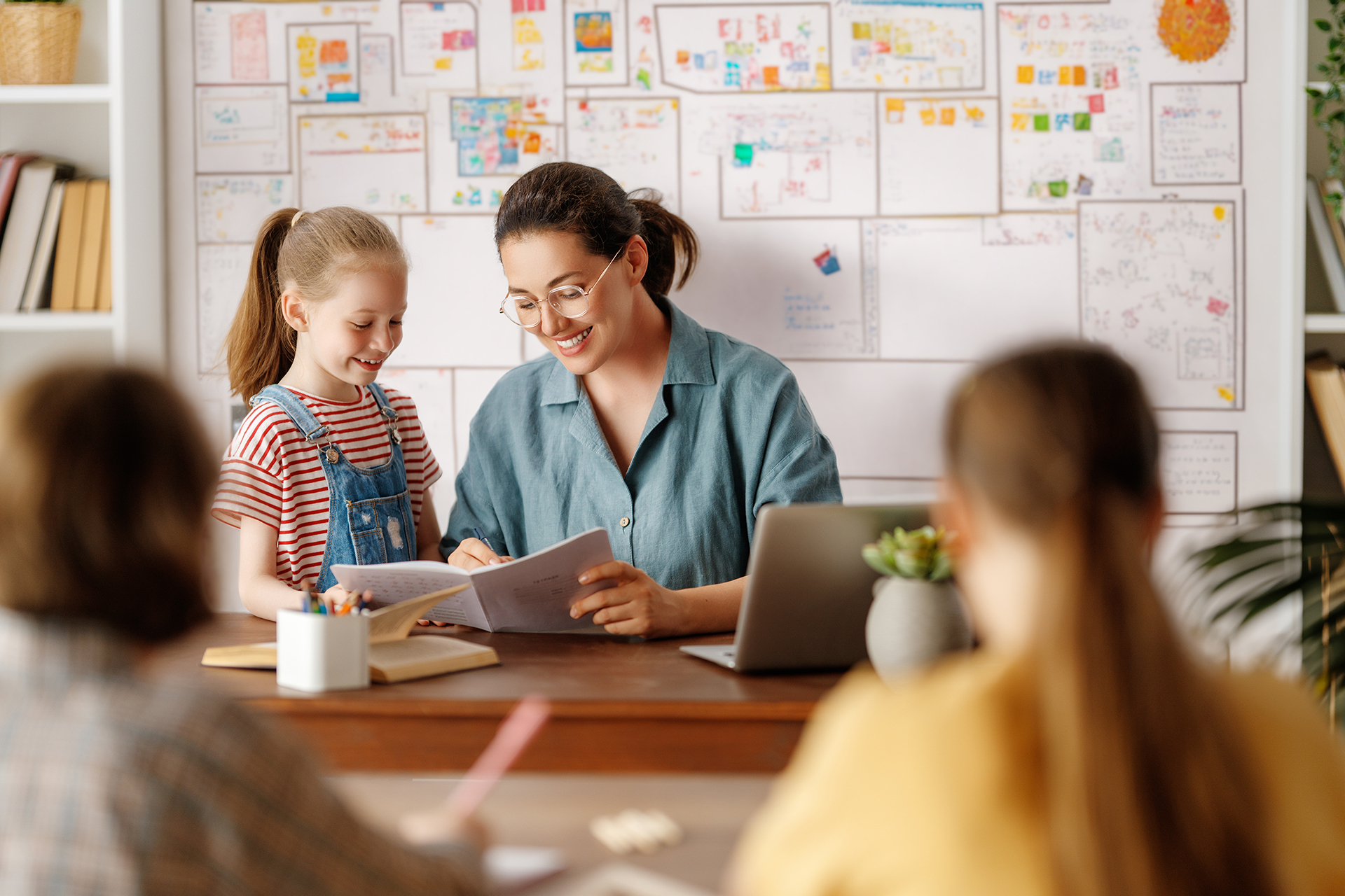 An female teacher seated at a desk reviews a notebook with a young student while other students sit nearby, with a wall of colorful drawings and a laptop visible in a classroom setting.