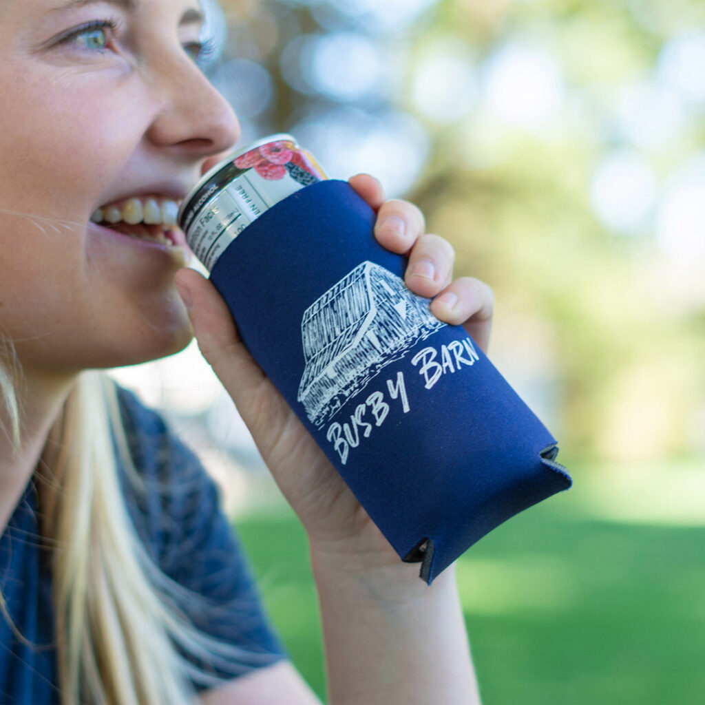 Close-up of a person drinking from a canned beverage inside a navy koozie printed with a barn illustration and the text “Busby Barn,” with a softly blurred green outdoor background.
