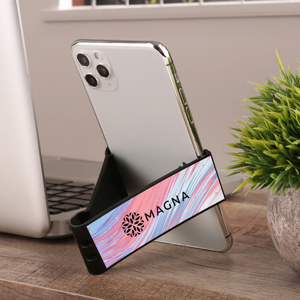 Smartphone propped on a compact black stand with a branded nameplate, placed on a desk next to an open laptop and a small potted plant.