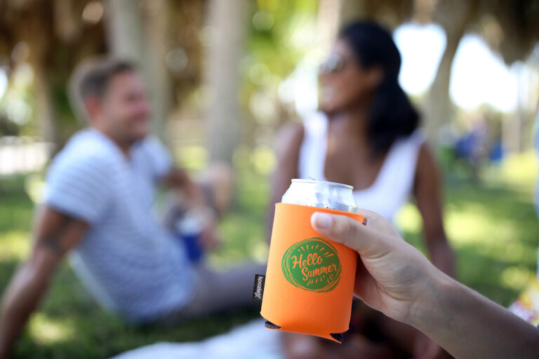 Hand holding a beverage can wrapped in a bright orange koozie with the text “Hello Summer,” in sharp focus, with two people sitting outdoors on grass and trees blurred in the background.