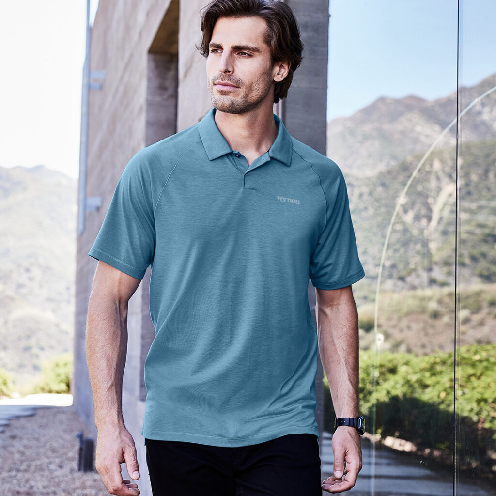 Person wearing a teal performance polo with a subtle tonal “Vernon” logo, walking in a modern outdoor setting with mountains in the background, highlighting refined, minimalist branded apparel.