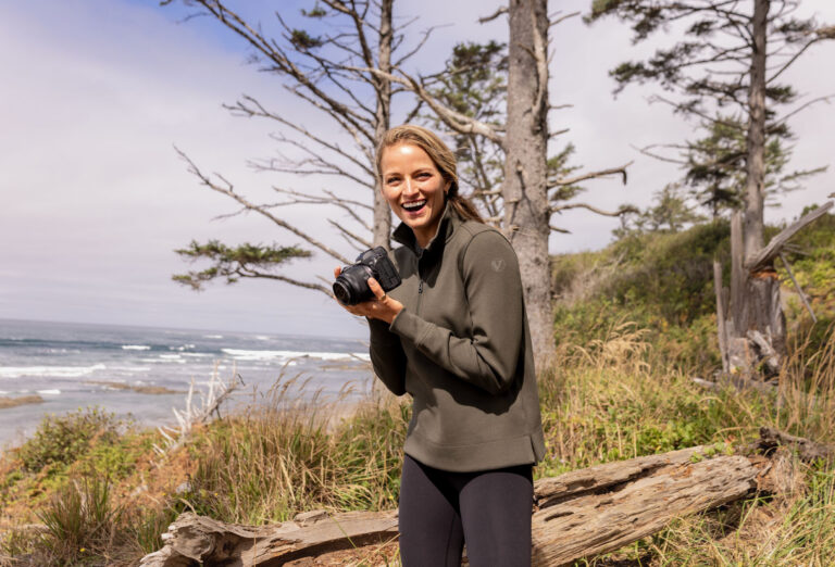 Person holding a camera on a coastal bluff, wearing a neutral-toned performance pullover with subtle tonal logo, illustrating understated, premium-style branded apparel in a natural outdoor setting.