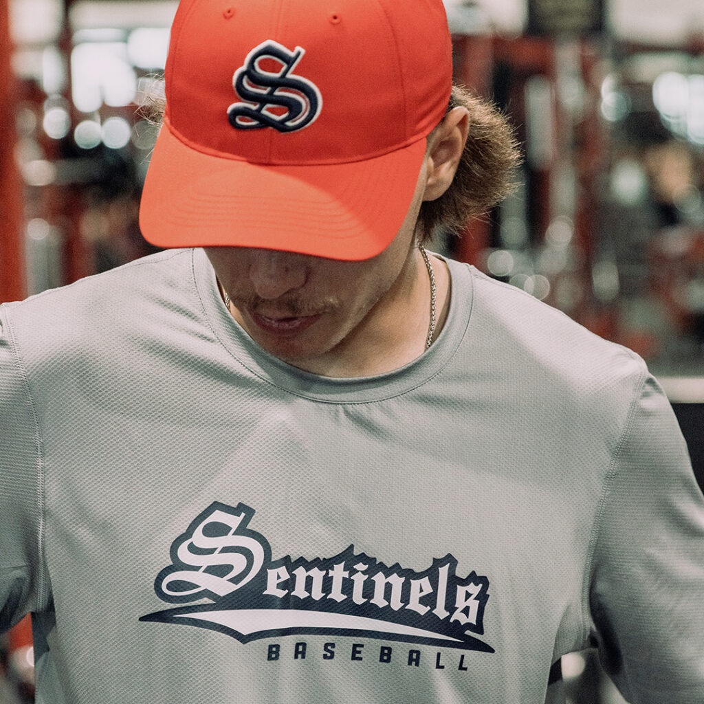 Close-up of a person in a gym wearing a red cap with an “S” logo and a gray “Sentinels Baseball” performance shirt, with weight equipment blurred in the background.