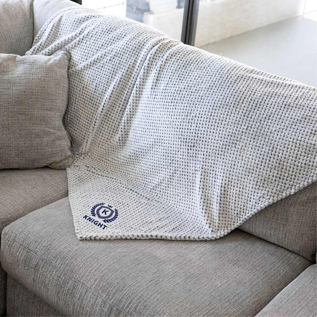 Soft, neutral-toned knit throw blanket with subtle embroidered “Knight” logo draped over a light gray sofa in a bright, modern living room, conveying understated luxury and minimal branding.