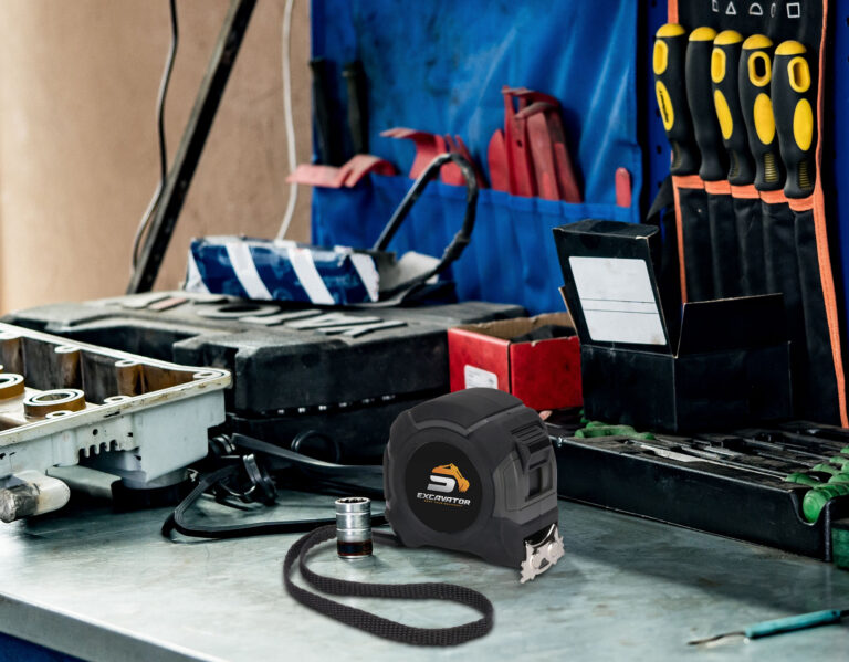 Branded black tape measure with custom excavator logo displayed on a mechanic’s workbench surrounded by hand tools and tool storage in a workshop.