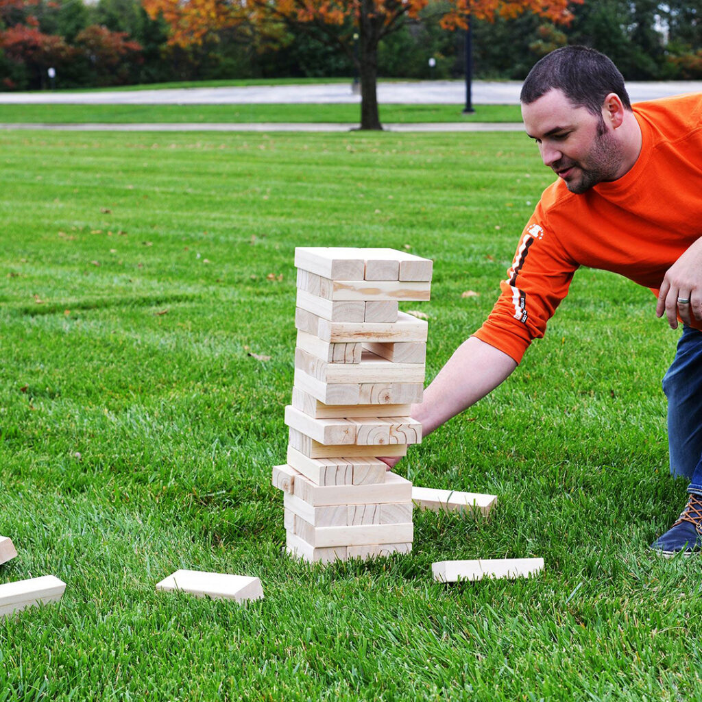 Person playing a giant wooden block stacking game on a grassy field, showcasing outdoor entertainment and tailgating event activities.