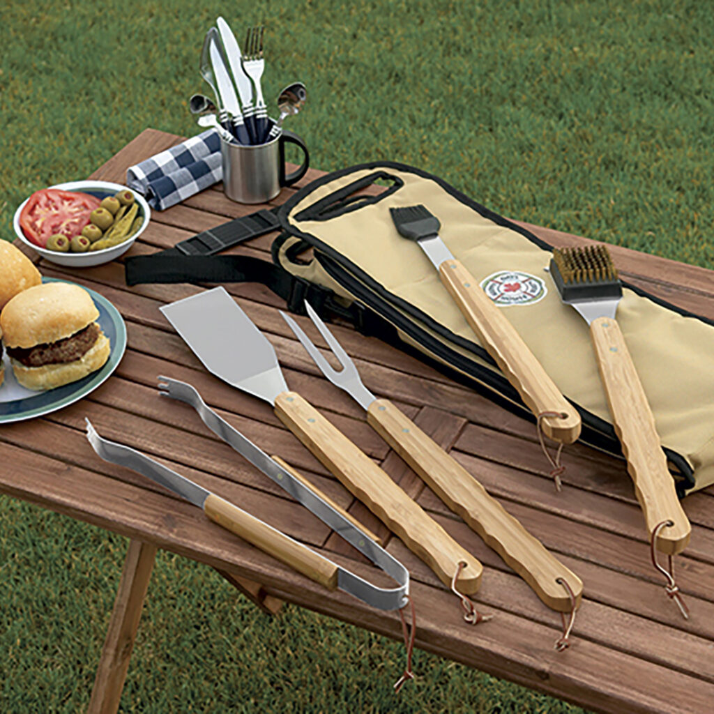 Branded barbecue tool set with spatula, tongs, grill fork, and basting brush displayed on a picnic table beside burgers and food prep items for tailgating.