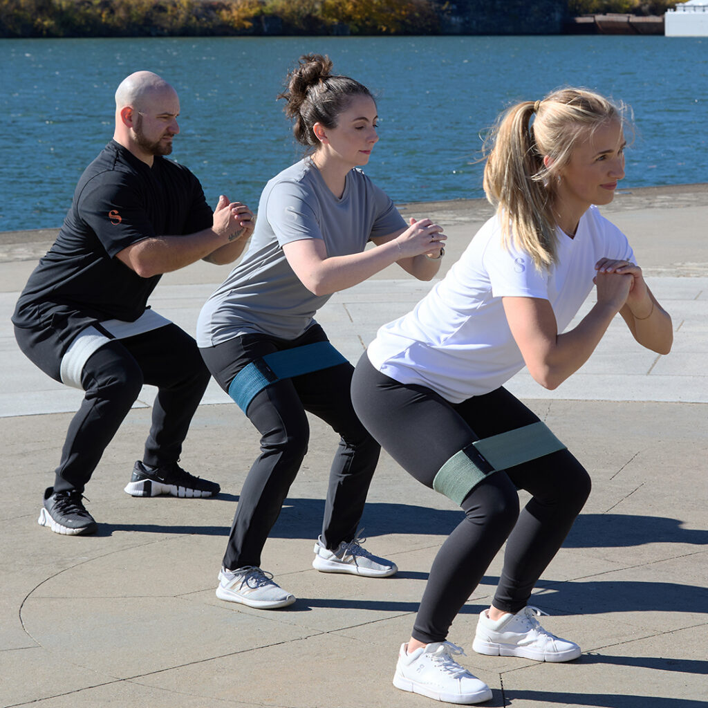 Three people performing resistance band squats outdoors near a waterfront as part of an active wellness routine.