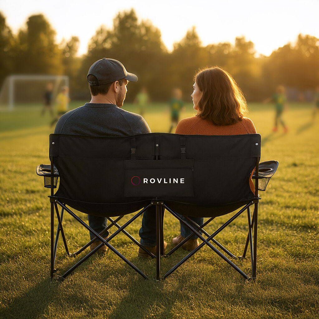 Custom-branded double folding camp chair set up on a grassy sports field, ideal for tailgating, outdoor events, and game-day seating.