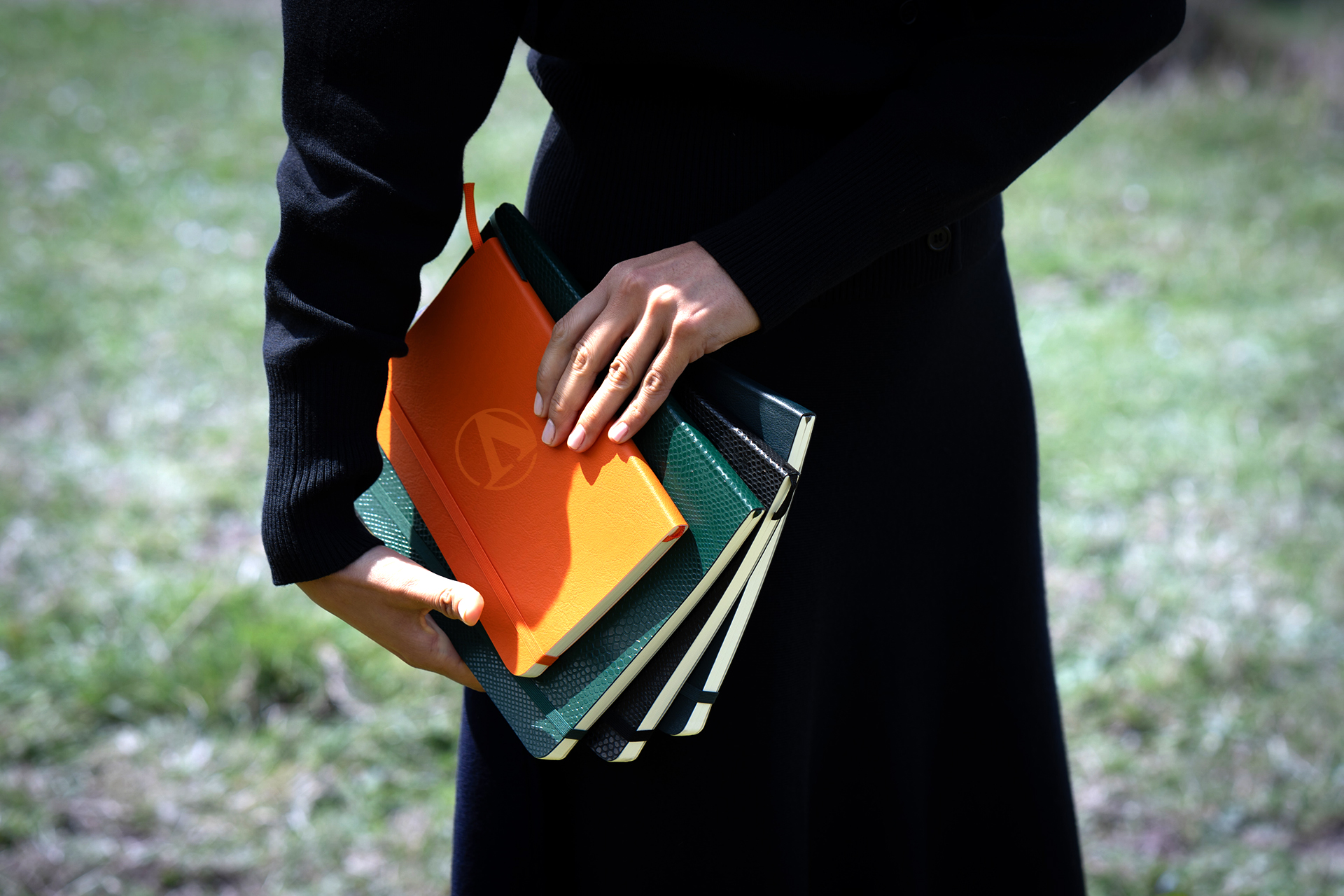 Close-up of a person carrying multiple textured hardcover notebooks, featuring an orange notebook with a debossed logo on the cover.