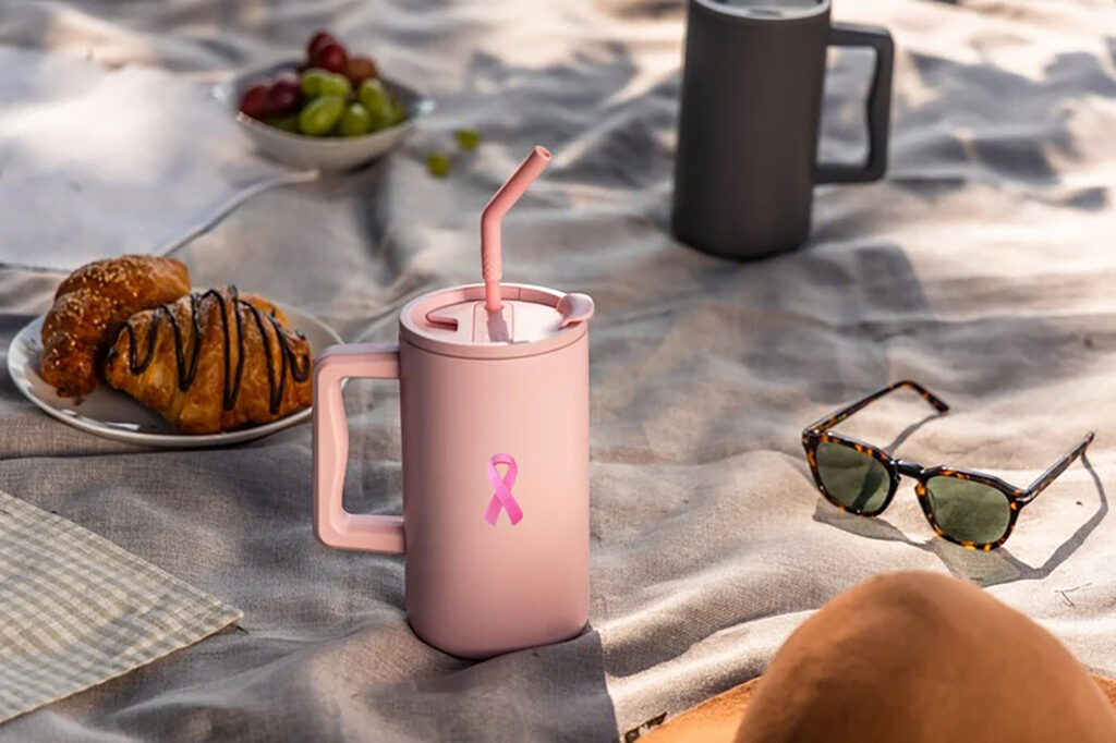 Pink straw tumbler with a breast cancer awareness ribbon logo on a picnic blanket.