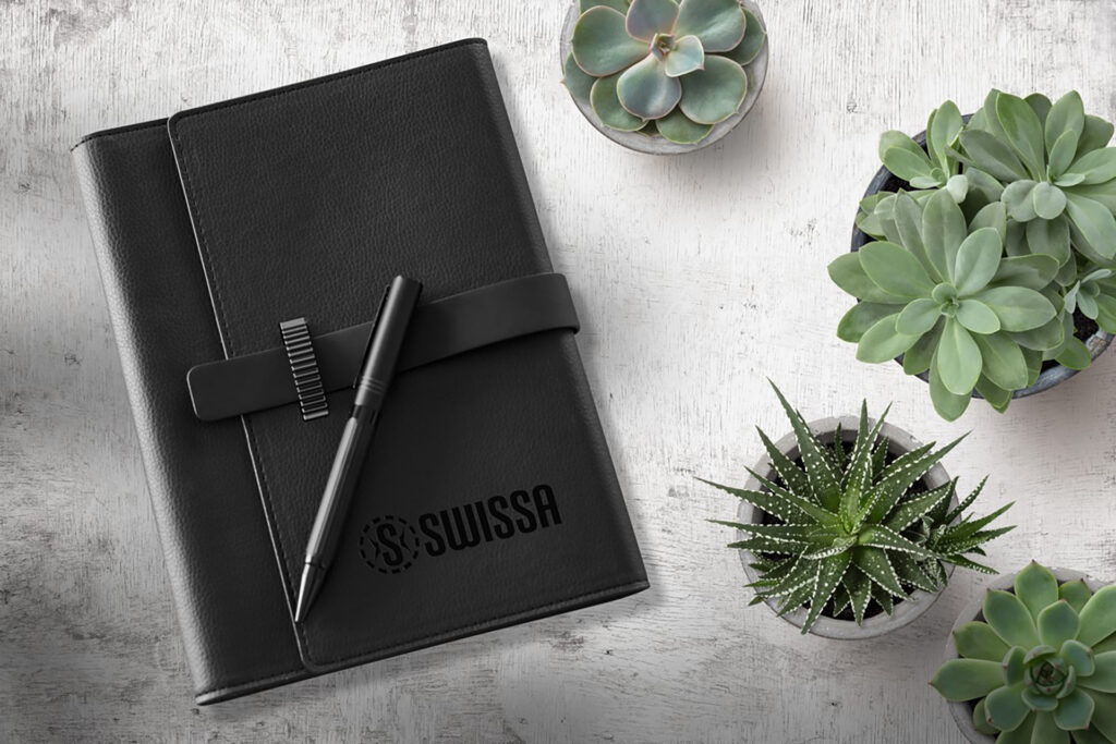 Black leather-bound branded journal with matching pen displayed on a light wood surface alongside potted succulents.
