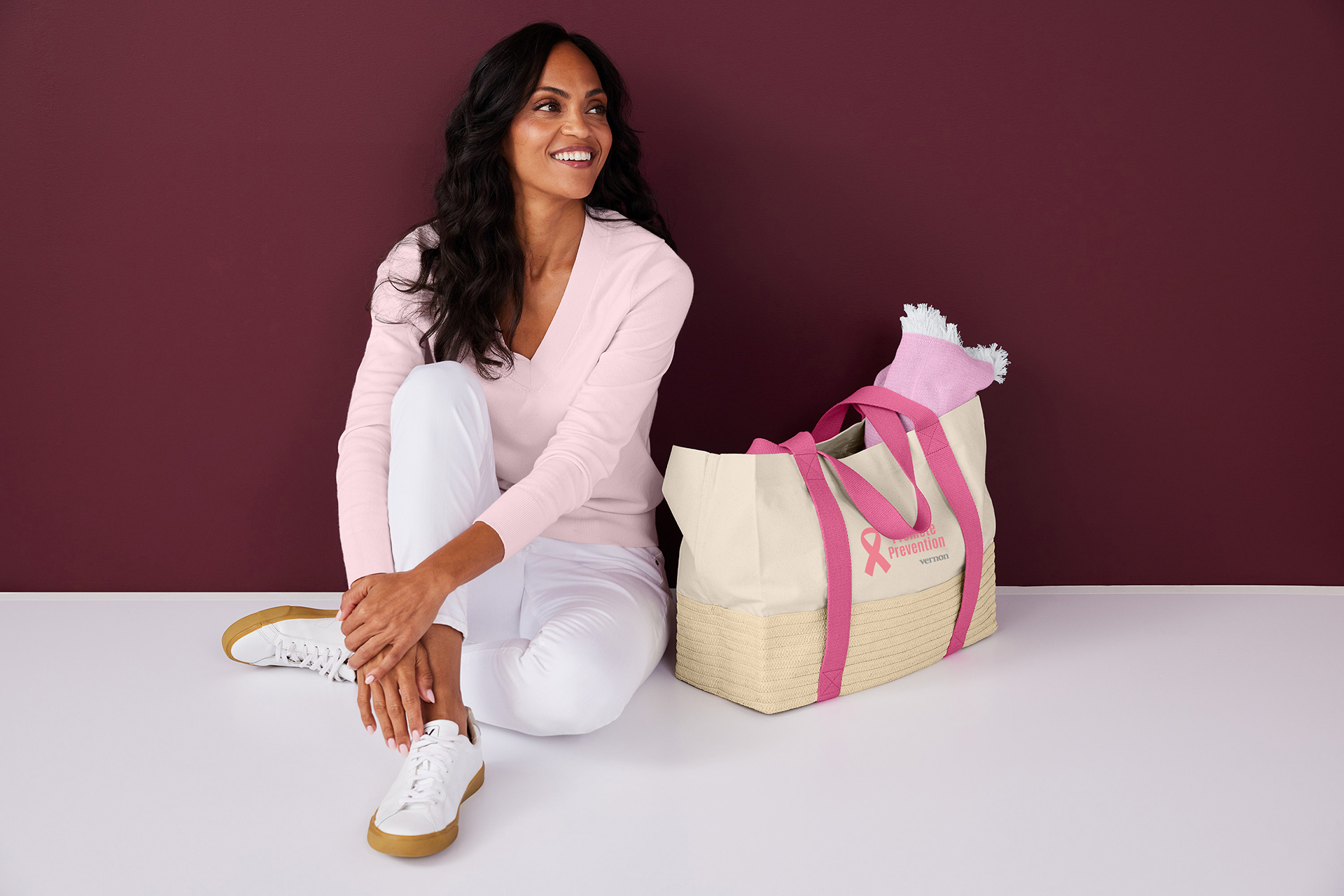 Person wearing a light pink sweater and white pants seated beside a promotional tote bag with pink handles and a breast cancer awareness ribbon logo, highlighting branded products for Breast Cancer Awareness Month.