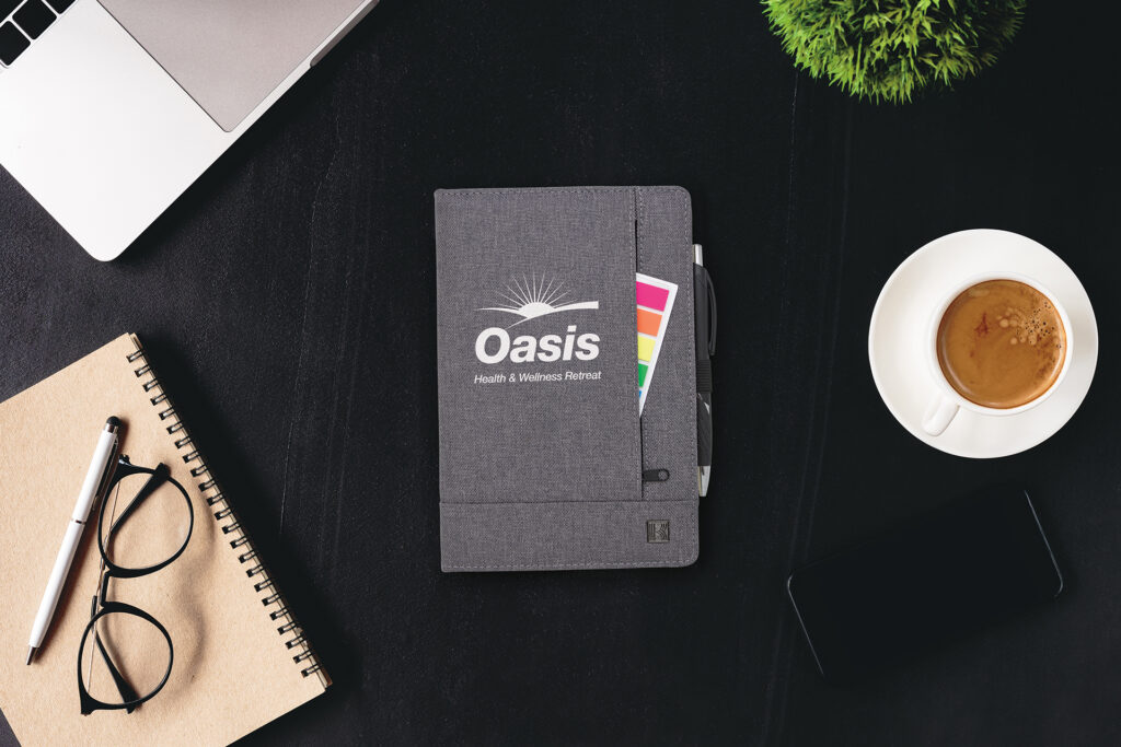 Branded gray notebook with logo, sticky note tabs and pen on a desk beside a laptop, coffee cup, eyeglasses and smartphone.
