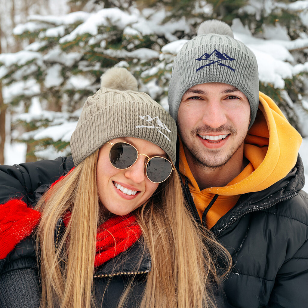 Two people wearing custom knit beanies in soft gray and warm neutral tones in a snowy outdoor setting, highlighting seasonless Autumn/Winter 2026 color trends for branded apparel and promotional merchandise.