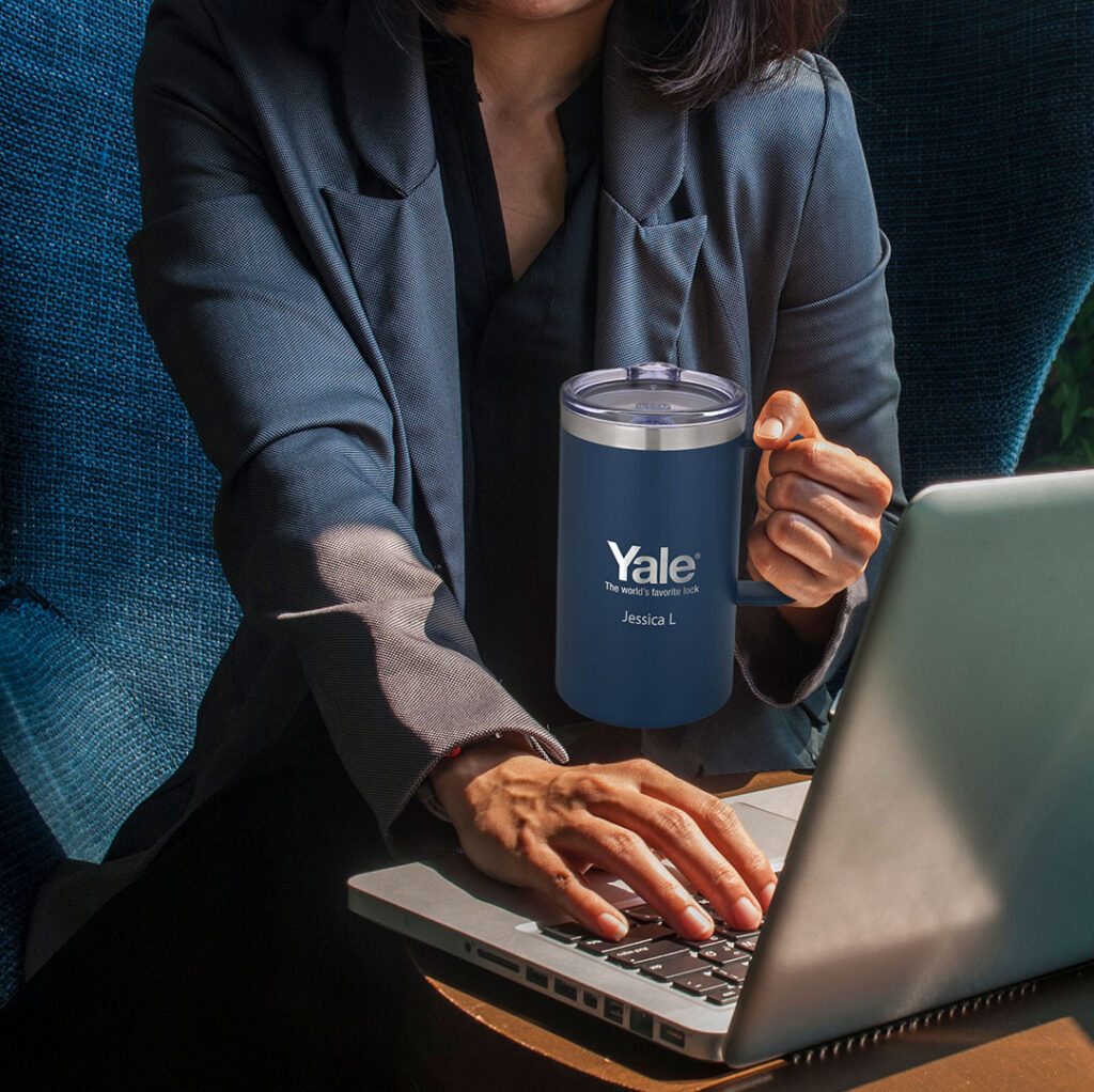 Custom insulated travel mug beside a laptop in a workspace, highlighting personalized drinkware as a popular promotional item for tech employees.