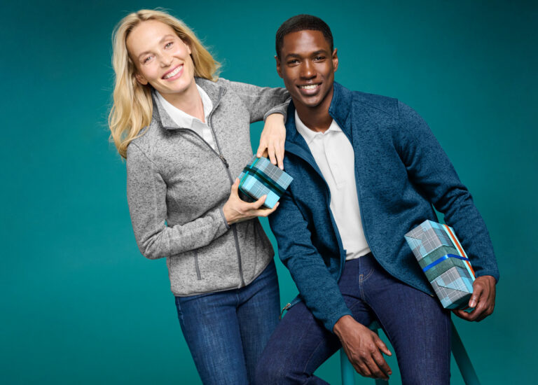 Two people wearing fleece jackets hold wrapped gift boxes, illustrating holiday gifting with branded merchandise.