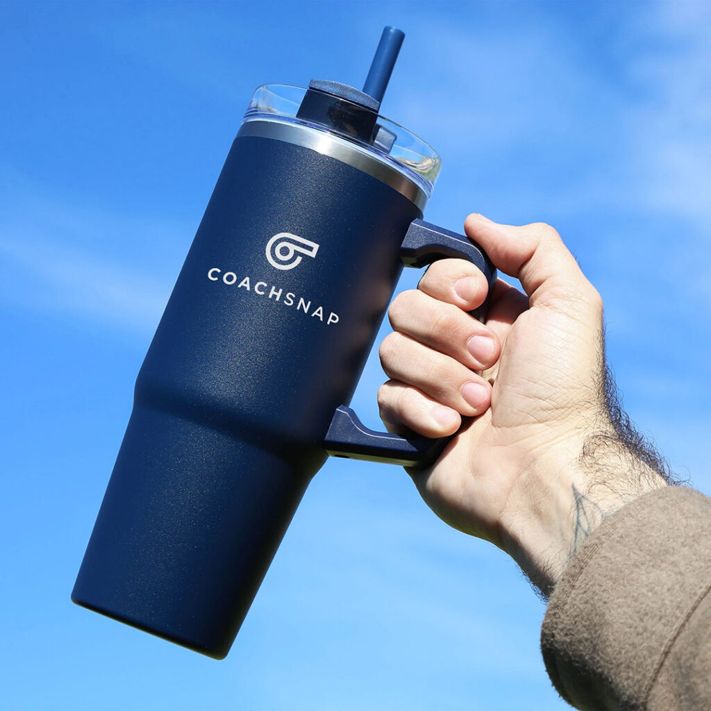 Branded insulated tumbler in a deep navy blue shade held against a clear blue sky, demonstrating how rich seasonless Autumn/Winter 2026 colors can create bold, functional promotional drinkware.