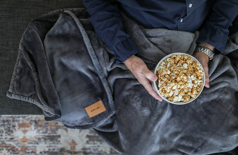 Close-up of a person relaxing on a couch with a luxury gray branded blanket and a bowl of popcorn, highlighting comfort, warmth, and the appeal of custom promotional blankets.