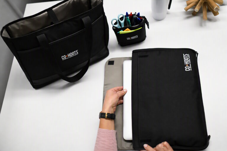 Black mobile office organizer sleeve holding a laptop on a white desk, with a matching black tote bag and desktop organizer nearby. The coordinated workspace accessories feature the CO+HOOTS logo, along with pens, scissors, sticky notes, and a white travel tumbler in a modern office setting.