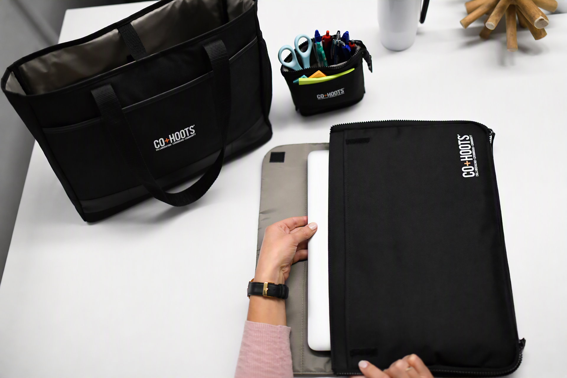 Black mobile office organizer sleeve holding a laptop on a white desk, with a matching black tote bag and desktop organizer nearby. The coordinated workspace accessories feature the CO+HOOTS logo, along with pens, scissors, sticky notes, and a white travel tumbler in a modern office setting.