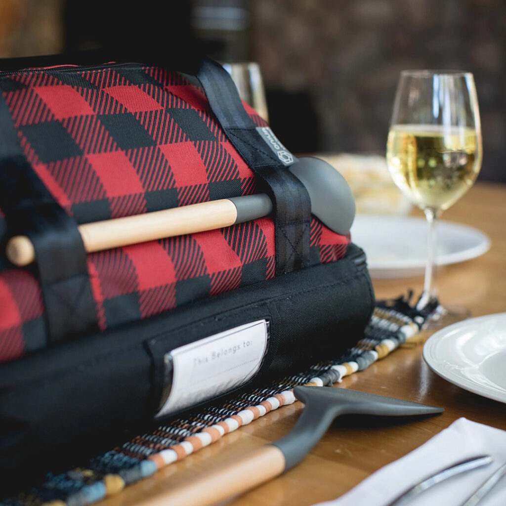 Red and black plaid insulated potluck bag with serving accessories displayed on a dining table.