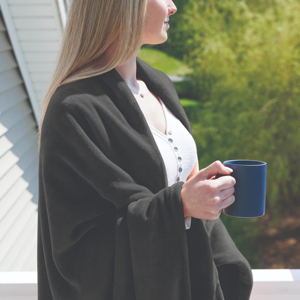 Person wrapped in a soft blanket while holding a coffee mug outdoors, representing a cozy lifestyle and a gift designed for comfort and relaxation.