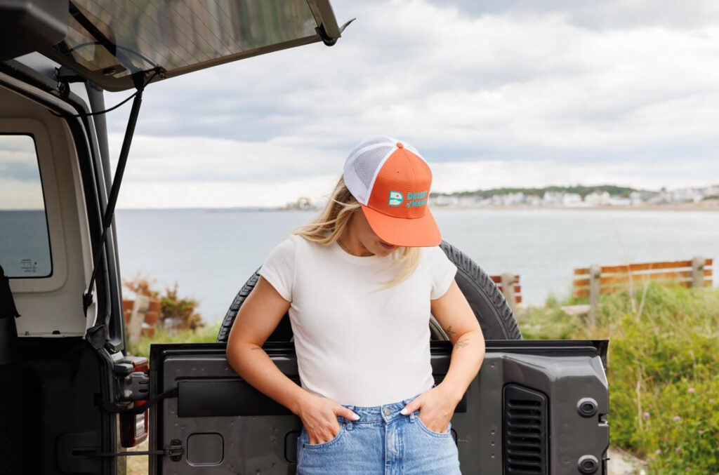 Branded trucker cap featured at the open tailgate of an SUV overlooking a waterfront.