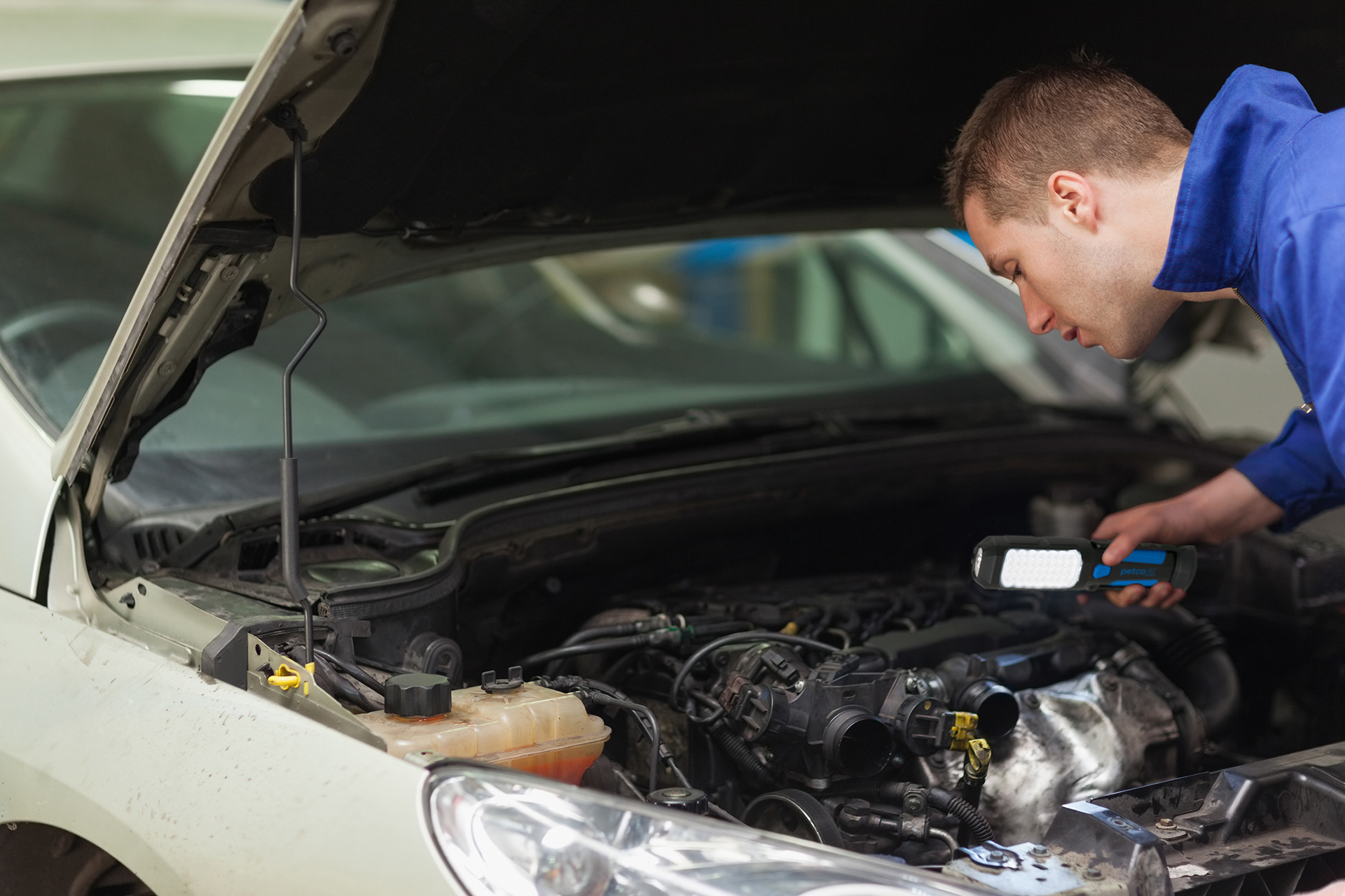 Person inspecting a vehicle engine with a handheld work light under an open hood in an automotive service setting, illustrating fleet maintenance, transportation reliability and vehicle upkeep.